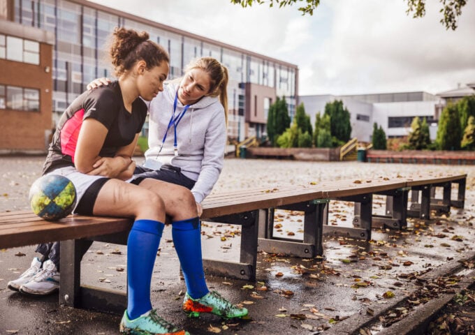 Young rugby player sitting on a bench outside a school with their head down looking sad next to rugby ball with their coaches arm over them trying to comfort them
