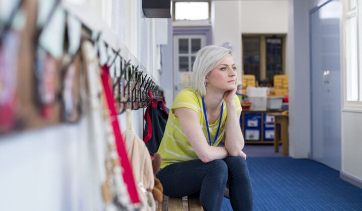 Young, female teacher sitting on a bench in the cloakroom of school. She is looking pensively out of the window.