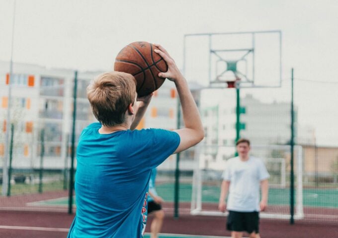 Young British basketball player wearing a blue shirt shooting basketball shot with team mate wearing white shirt in the guarding the net