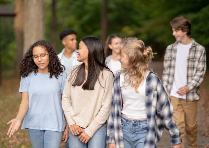A small group of teenagers walk casually outside on a fall day together as they laugh and talk amongst themselves. They are each dressed casually in warm layers and enjoying hanging out together.