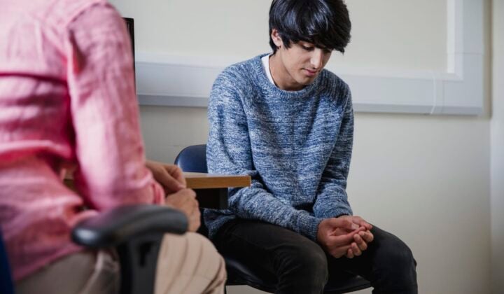 Young boy sitting on chair looking down with hands together whilst talking to person about their mental health