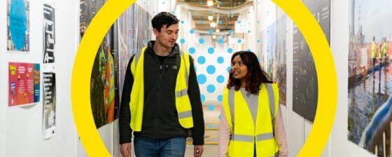 Two people walking along a corridor at work with high vis jackets